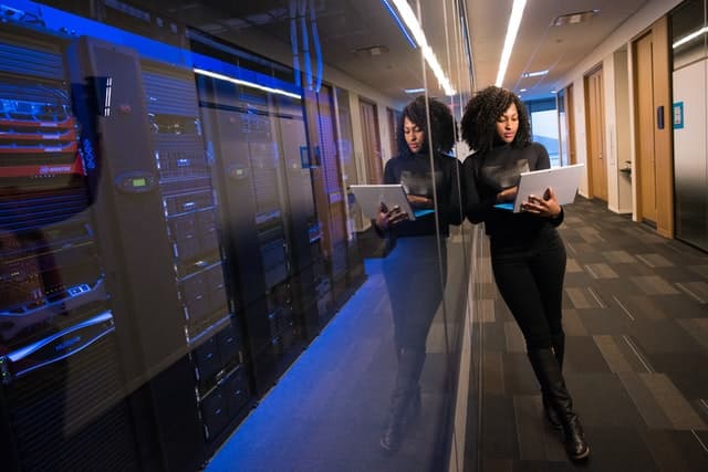 A lady stands next to website hosting servers, monitoring them using a laptop